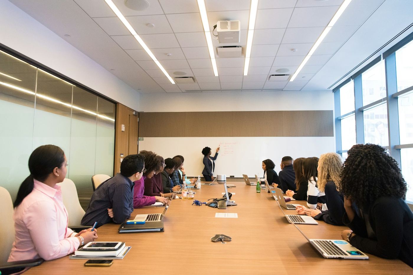 A large team seated around a boardroom table watching a colleague work through a whiteboard