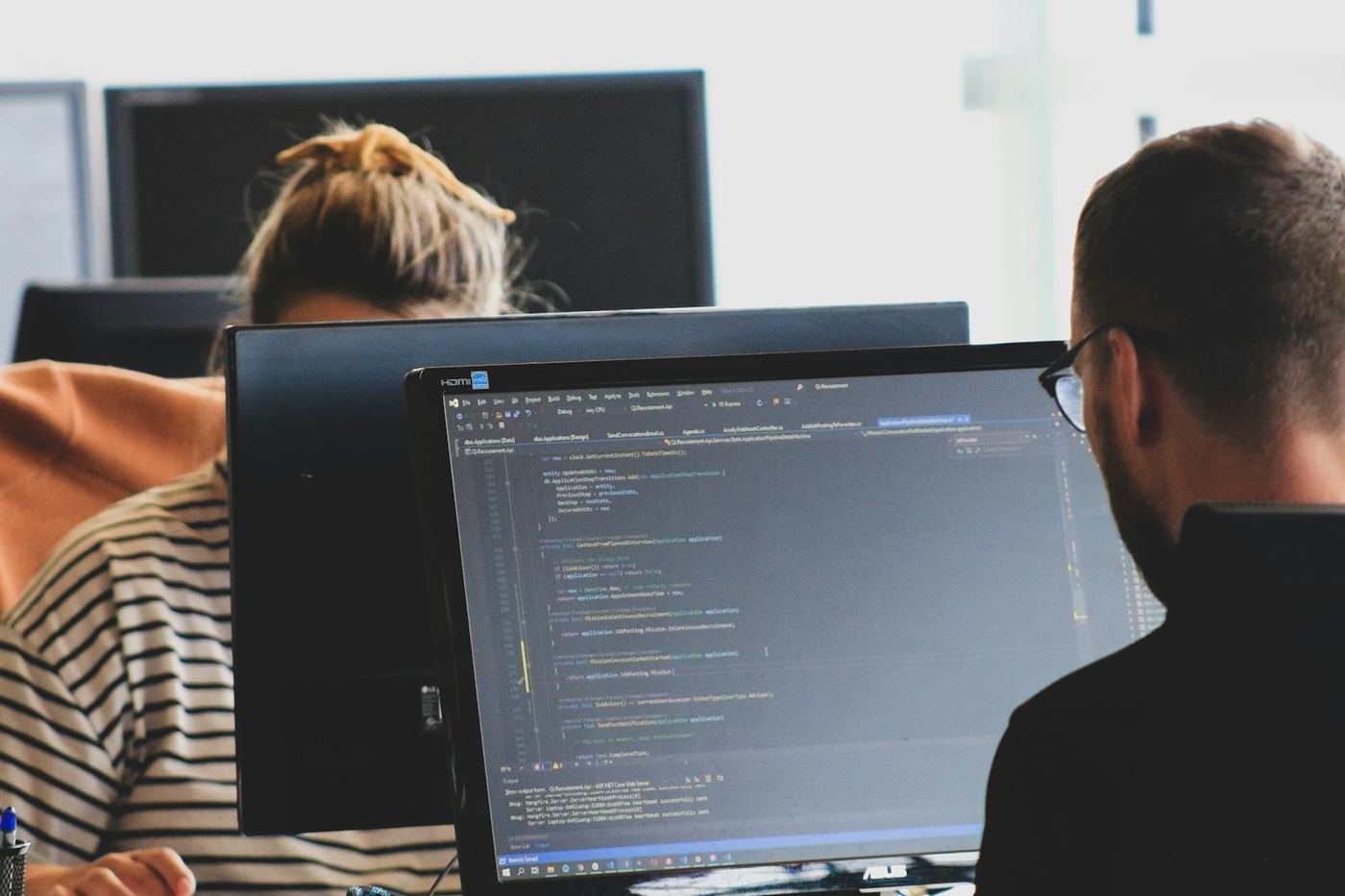Two developers working at adjacent monitors, one screen showing application code