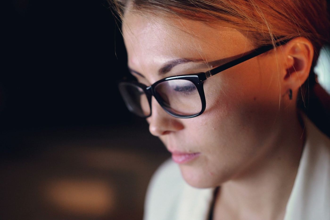 A woman in glasses reading closely from a screen, lit by the display
