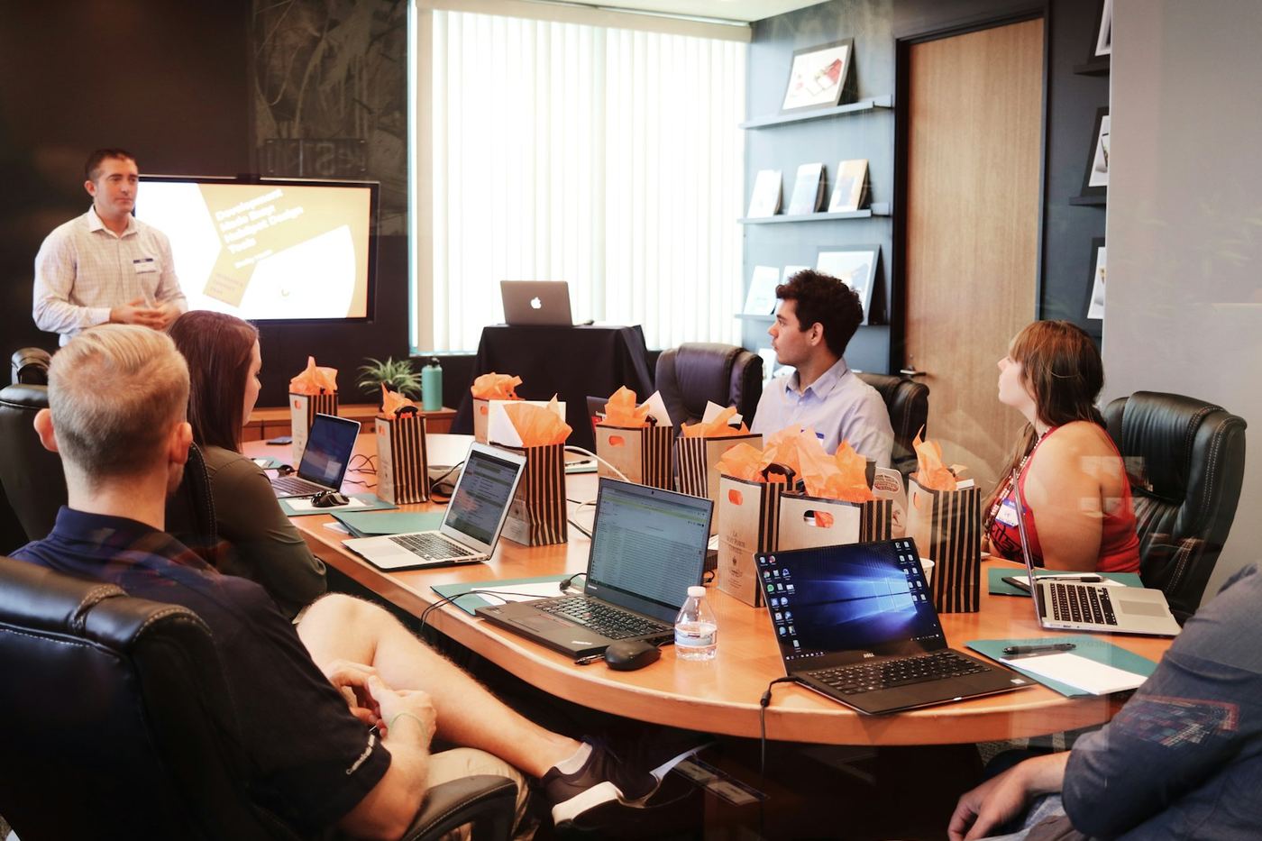 A facilitator presenting to a seated group working on laptops around a boardroom table
